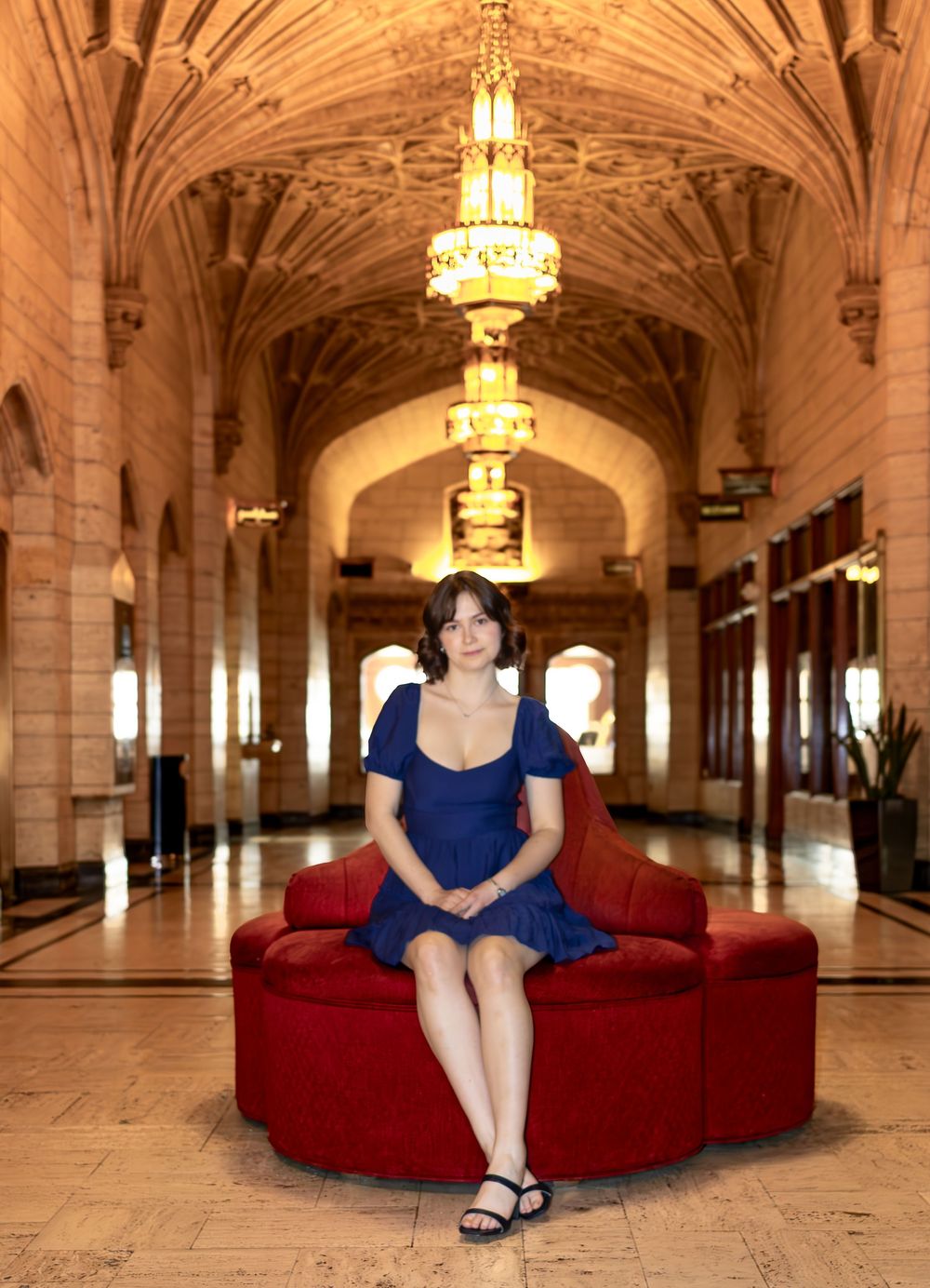 A person sits on a red velvet couch in an ornate hallway with arched ceilings and chandeliers.