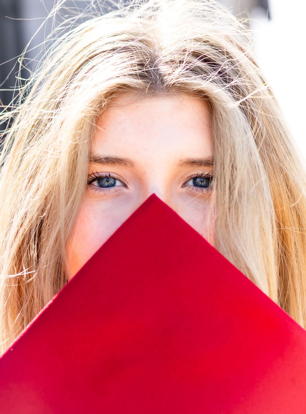 Blonde woman with blue eyes peeking over a red triangular shape covering her face.