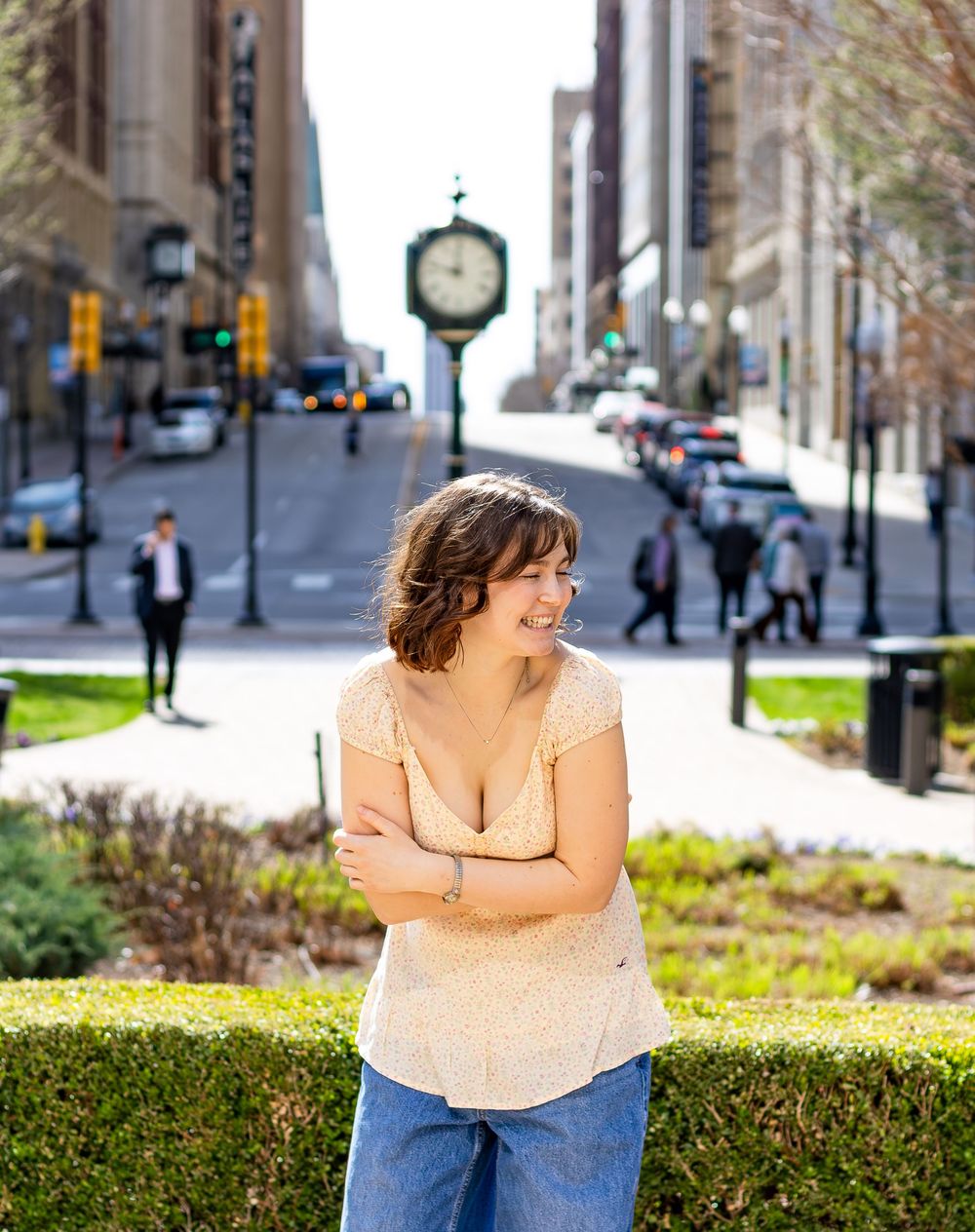 A person in a peach colored top and jeans sits casually on a stone ledge in an urban setting with trees and buildings.