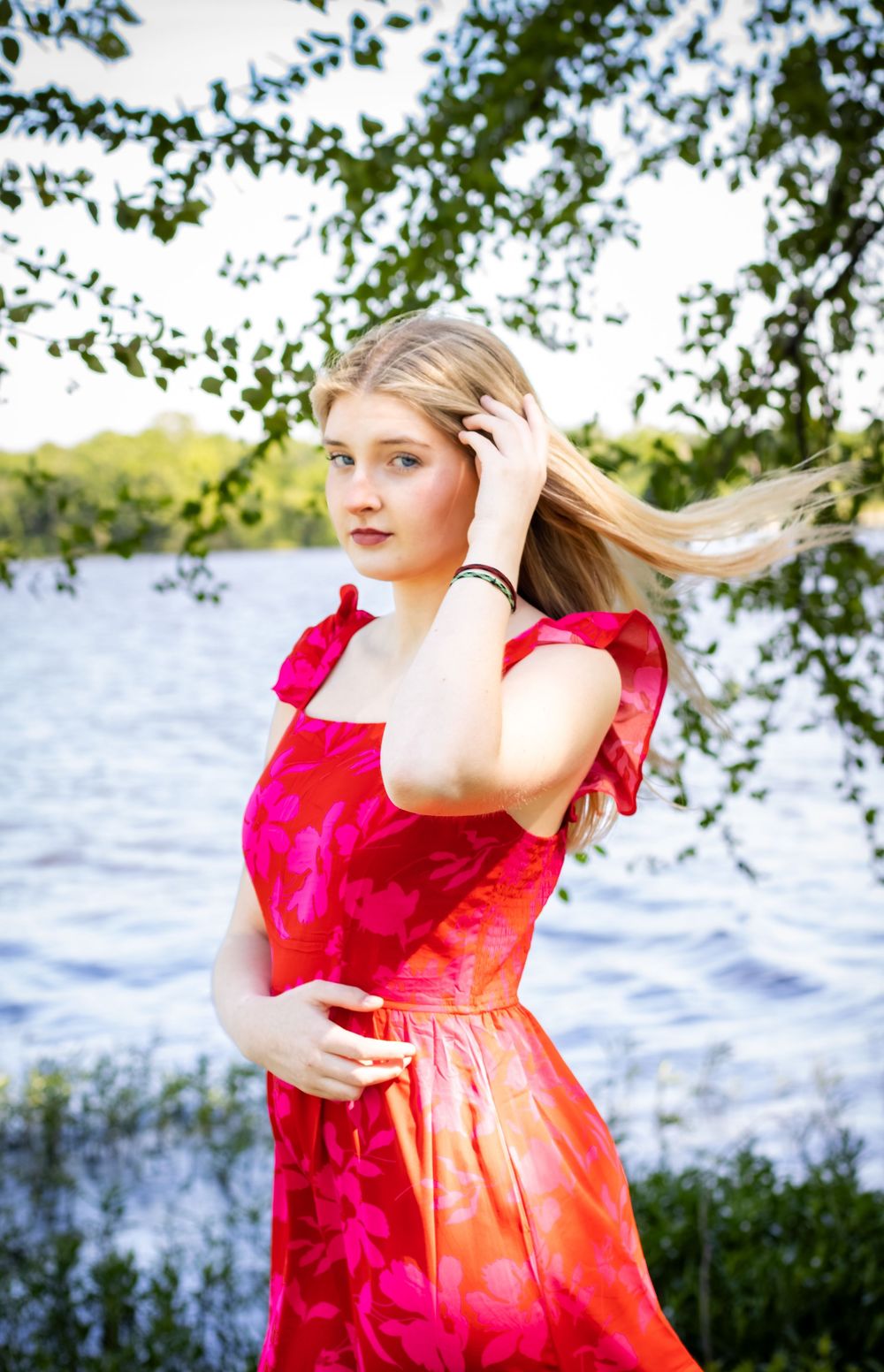 Young blonde woman in red floral dress poses gracefully by a lake with wind-blown hair under green trees.