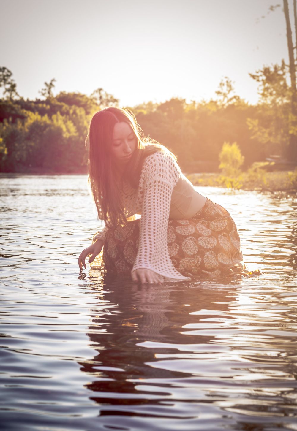 A young woman in a crochet top wades in a sunlit lake at golden hour, surrounded by lush green trees.