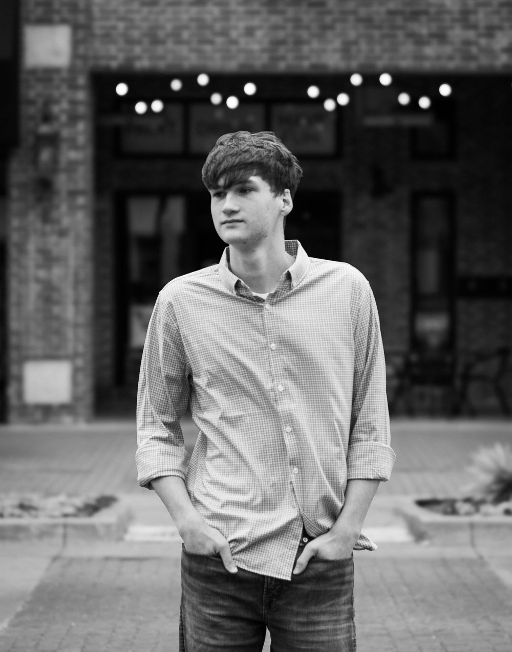 Young man in a button-up shirt stands outdoors with hands in pockets, string lights and brick building in background.