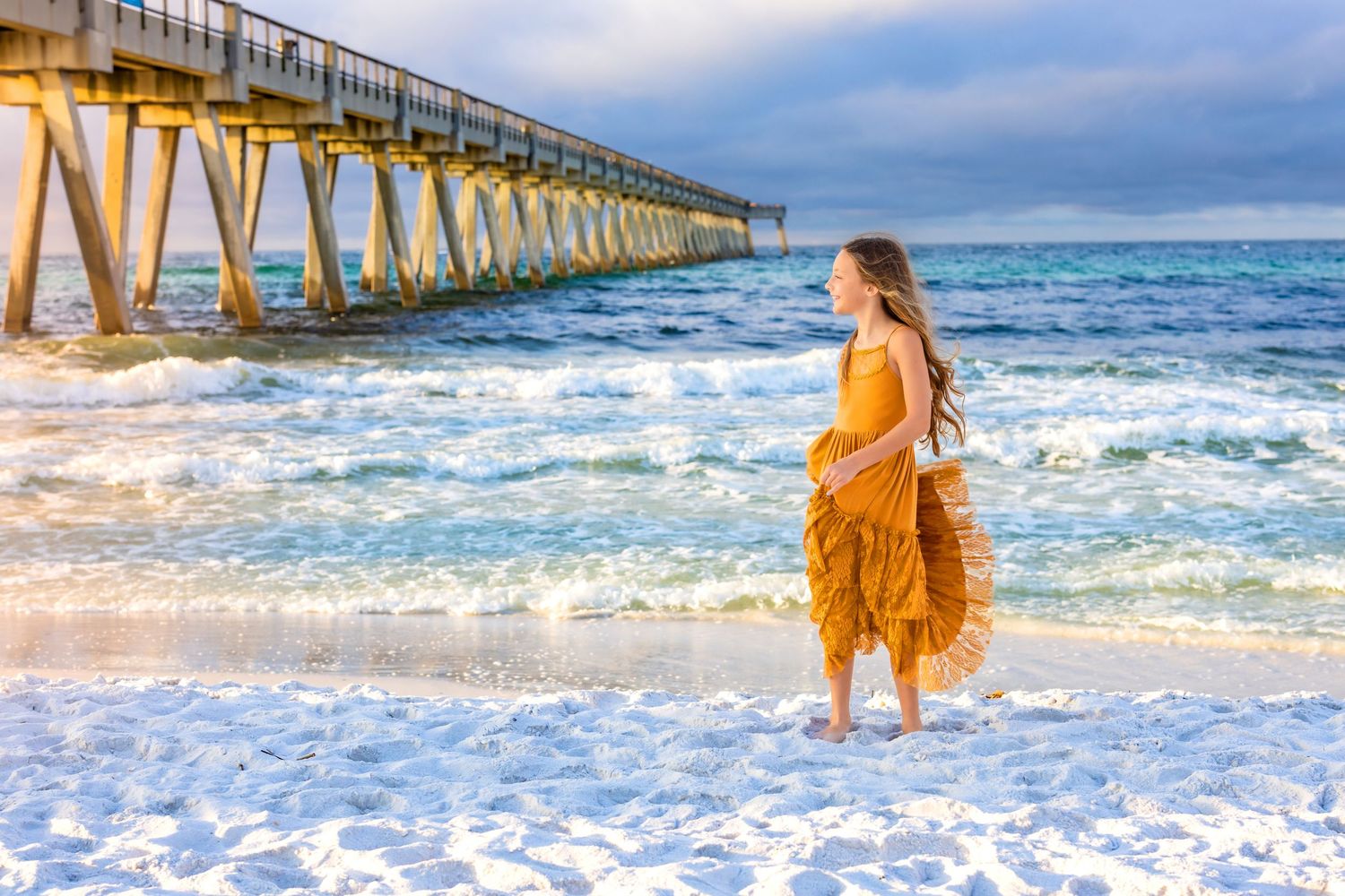 A person in a flowing yellow dress stands by the ocean waves near a wooden pier at sunset.
