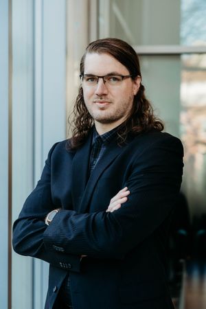 Professional man with long hair and glasses in a dark blazer, arms crossed, posing confidently near a window.