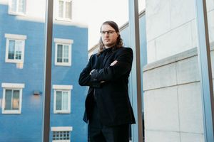 Man with long hair and glasses in black suit stands arms crossed near blue building exterior.