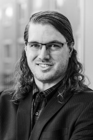 Black and white portrait of a young man with long curly hair and glasses wearing a suit jacket.
