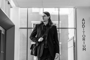 Man in black suit with long hair carrying a bag outside an auditorium entrance in black and white.