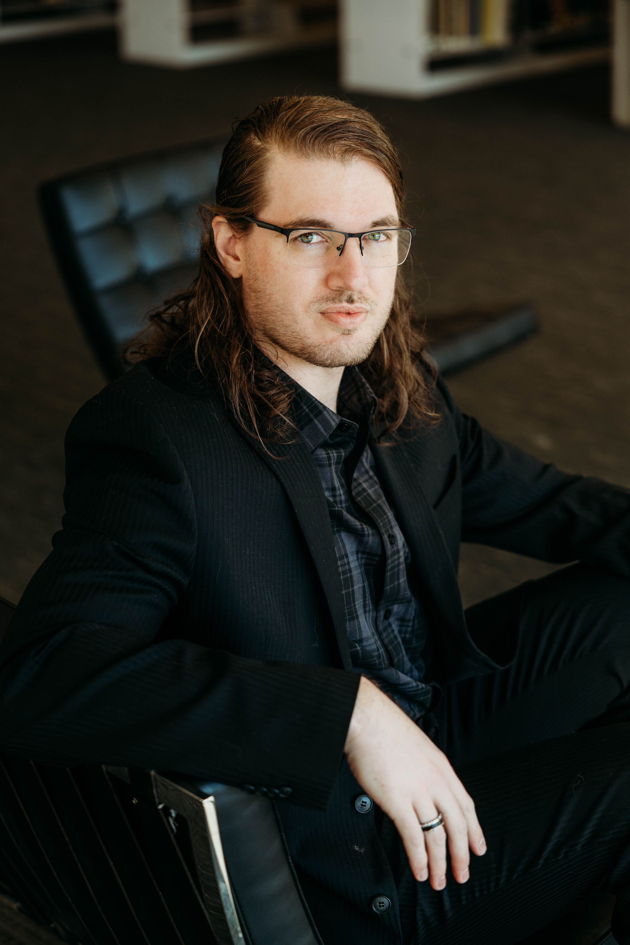 Young man with long hair and glasses in a black suit seated in a modern chair in a library.