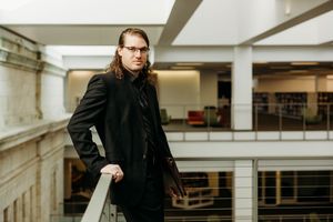 Young man with long hair and glasses in black suit leaning on railing in modern building interior.