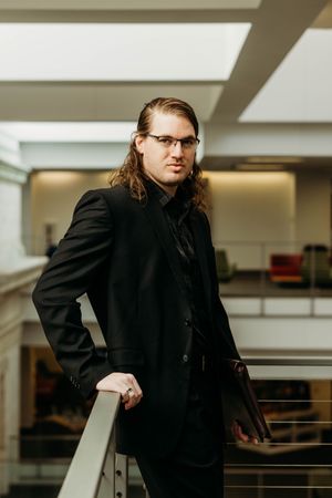 Stylish man with long hair wearing black suit and glasses posing confidently in modern atrium space.