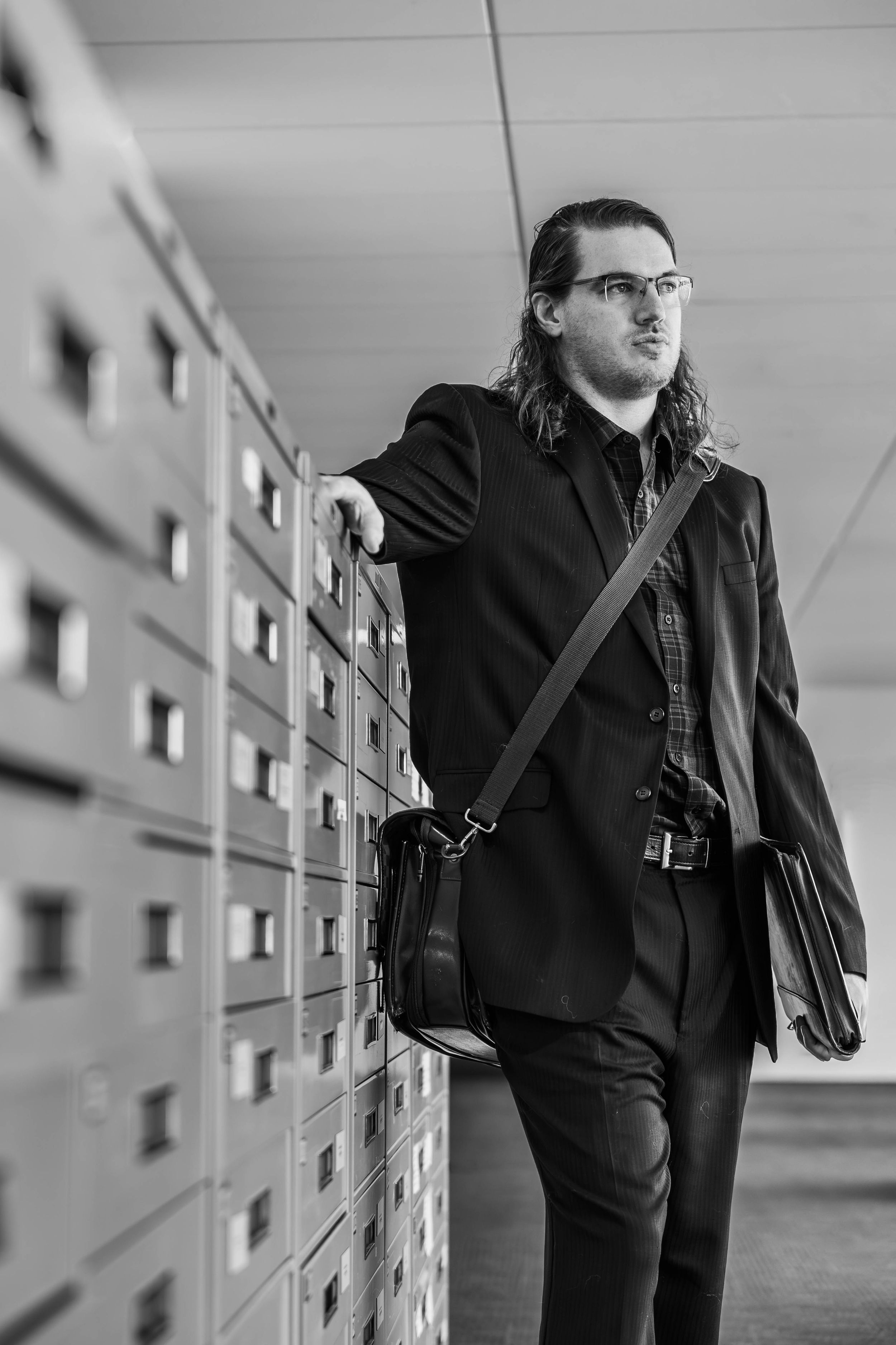 Black and white photo of a long-haired man in a suit with a messenger bag leaning against filing cabinets.