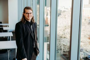 Young professional with long hair and glasses smiling in modern building lobby wearing black blazer.