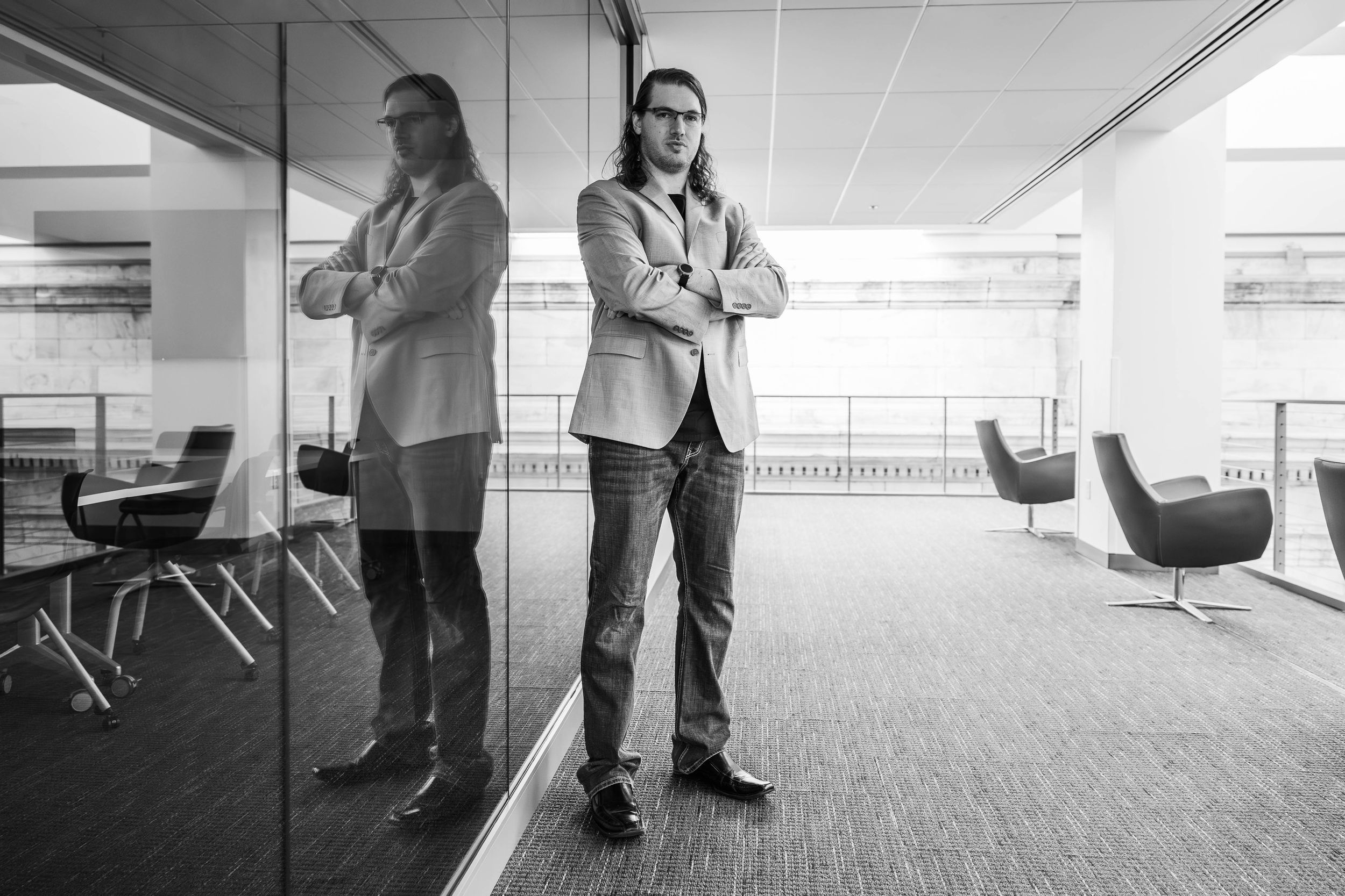Professional man with glasses stands confidently in modern office, arms crossed, reflected in glass wall.