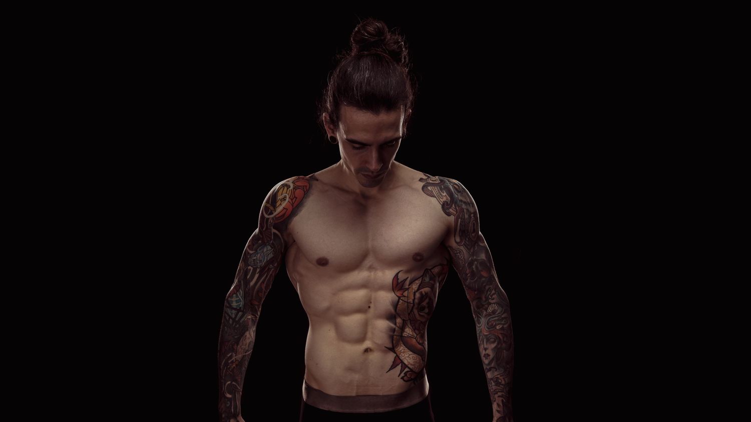 Shirtless tattooed man with sleeve tattoos and defined abs posing against a black background.