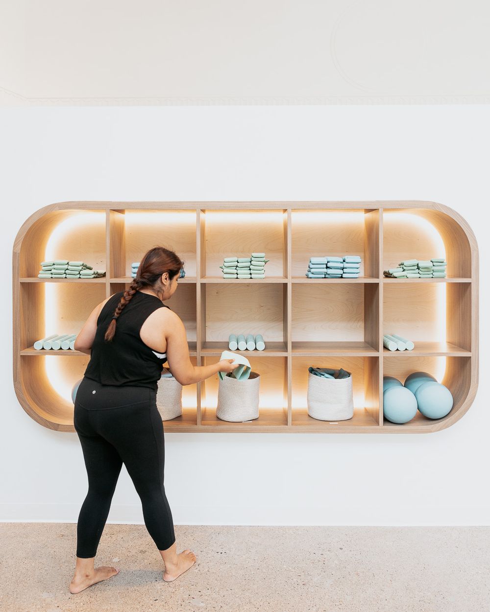 Woman in black activewear organizing mint green towels and accessories on a modern illuminated wooden shelf.