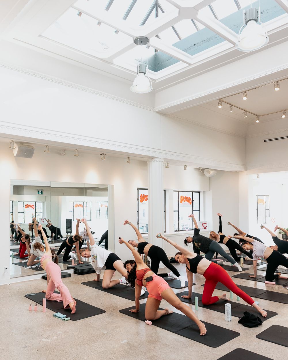 Large yoga class performing side stretch poses on black mats in a bright, modern studio with skylight ceiling.
