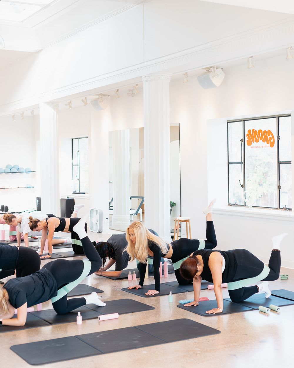 Group of women practicing yoga poses on black mats in a bright, modern studio with white columns and mirrors.