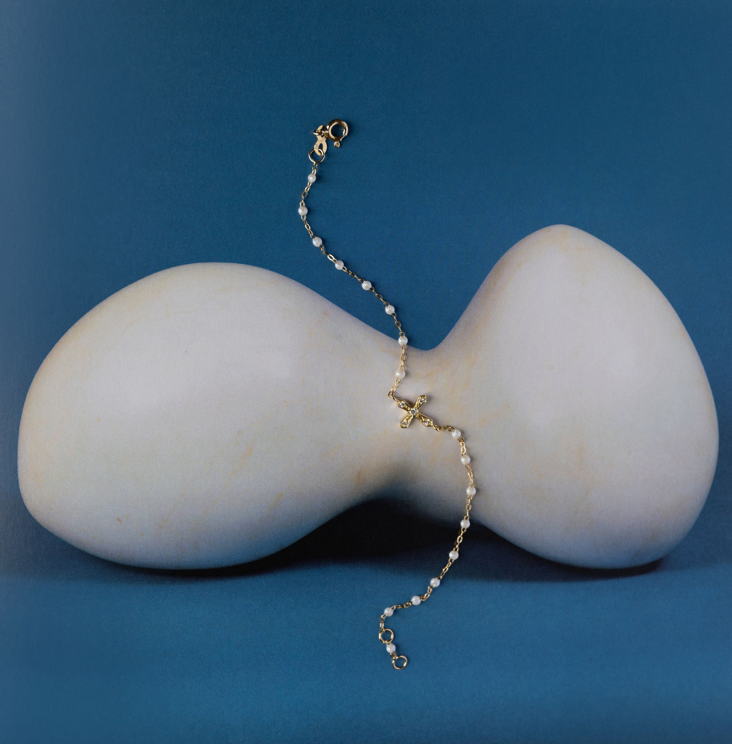 Delicate gold chain bracelet with pearl accents and cross charm displayed on white sculptural prop against blue background.