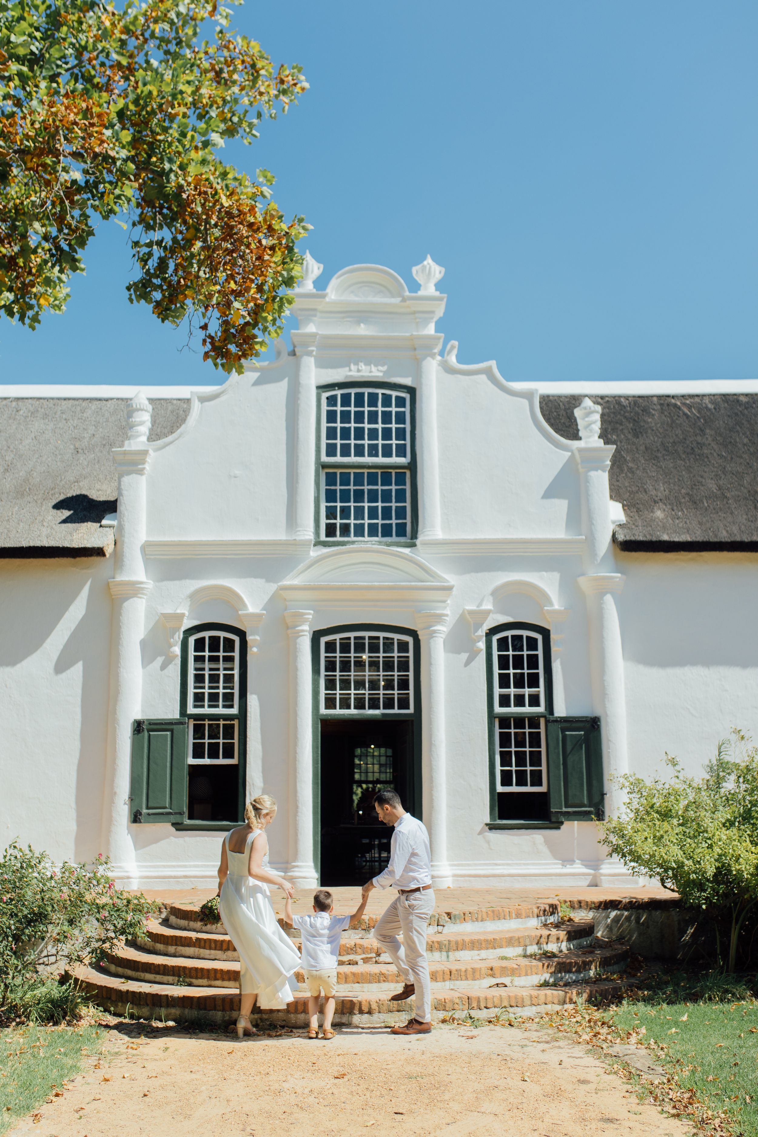 Couple with child on steps of white Cape Dutch manor house with green shutters and thatched roof under blue sky.
