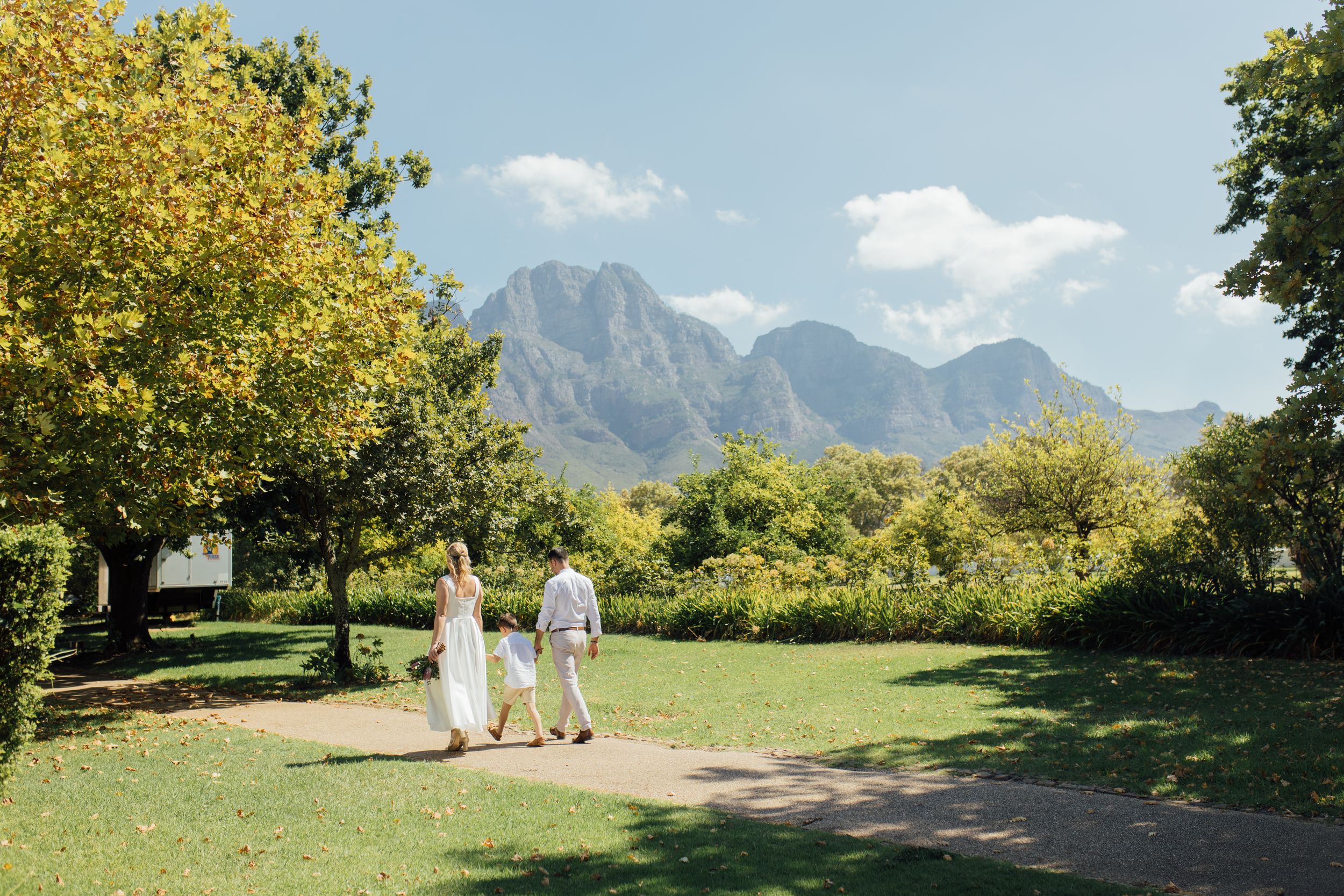 Couple walking hand-in-hand through lush garden with majestic mountain backdrop on sunny day.