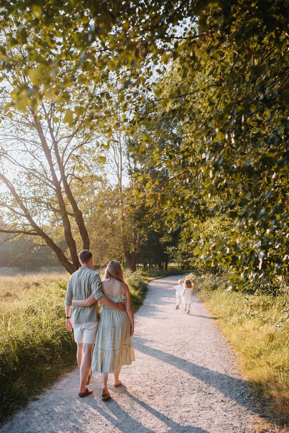 Family walking on a sunlit gravel path through lush green trees, parents embracing as child runs ahead.