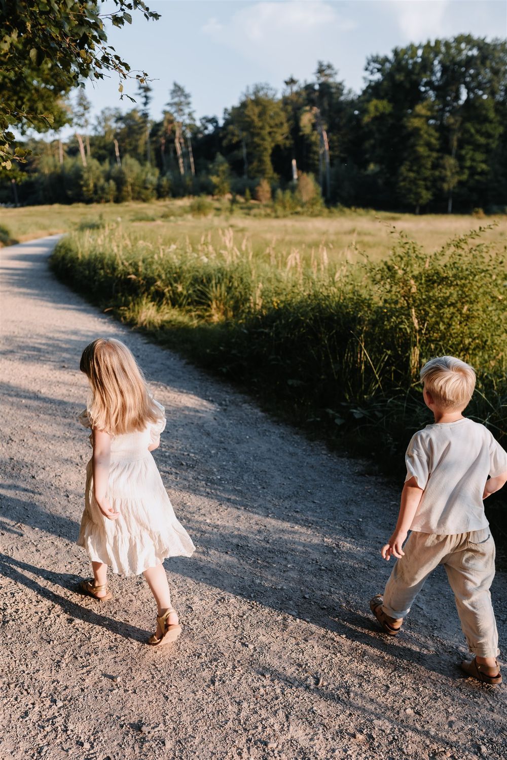 Two young children walk along a sunny gravel path surrounded by lush green fields and trees.