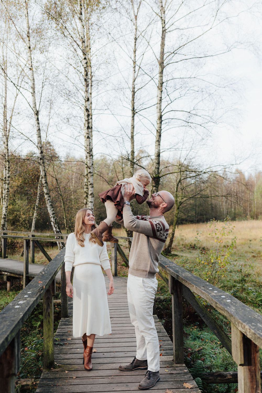 A happy family on a wooden bridge, with a man lifting a baby while a woman in white dress watches, surrounded by birch trees.