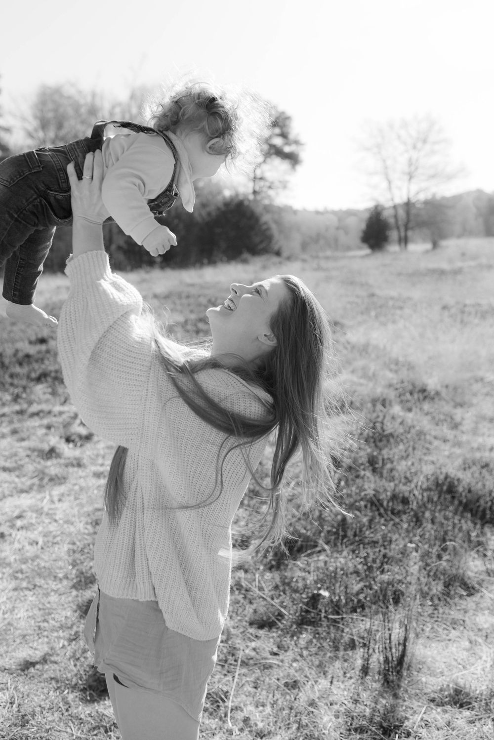 Black and white photo of a smiling woman lifting a happy baby overhead in a sunny outdoor field.