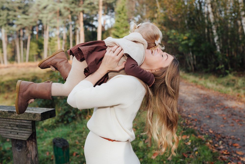 Mother lifting and kissing her baby outdoors on a forest path in autumn, both wearing warm brown and cream outfits.