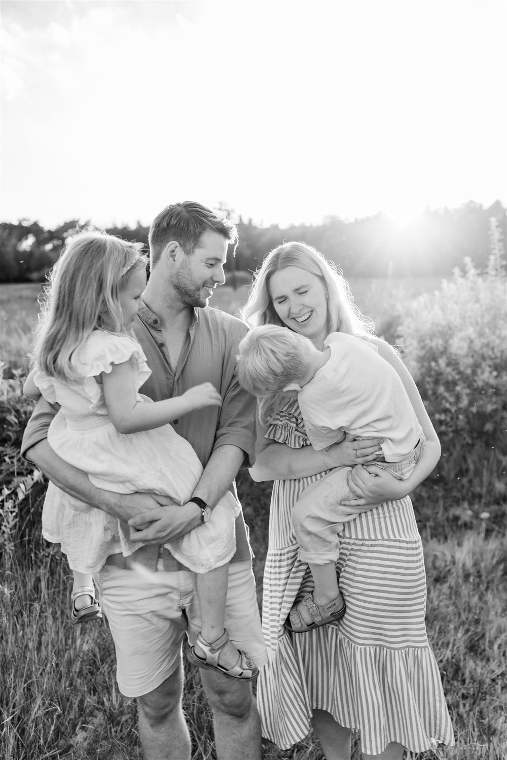 Black and white family photo of parents holding two young children laughing outdoors in a sunny field.
