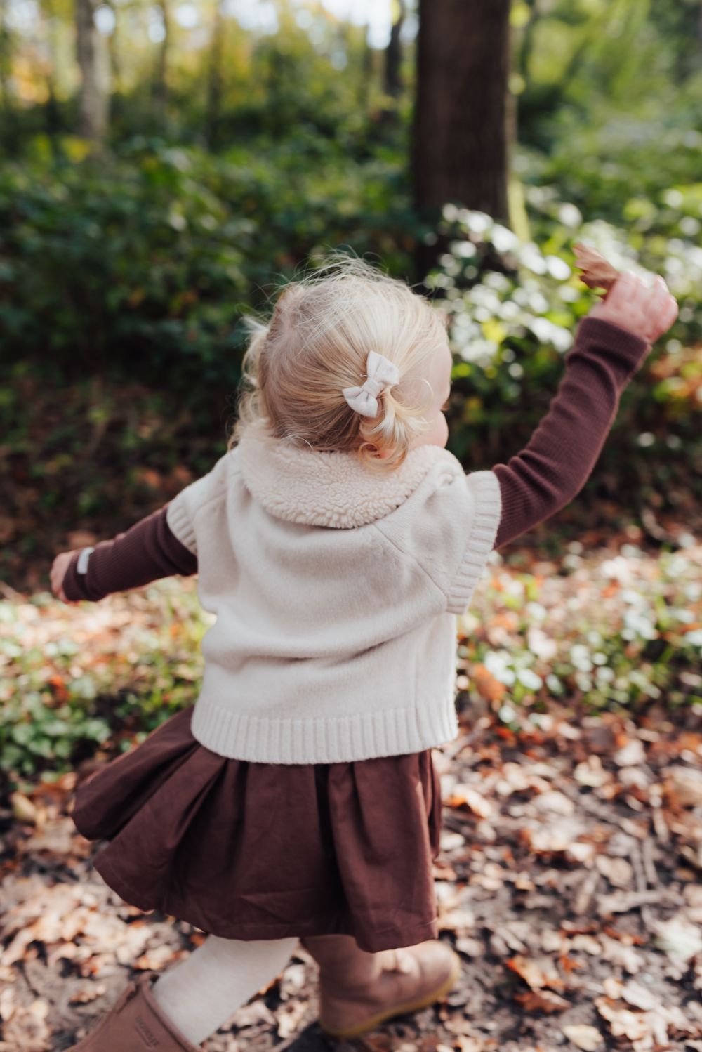 Toddler girl in cream sweater vest and burgundy skirt playing in autumn leaves in a lush green forest.