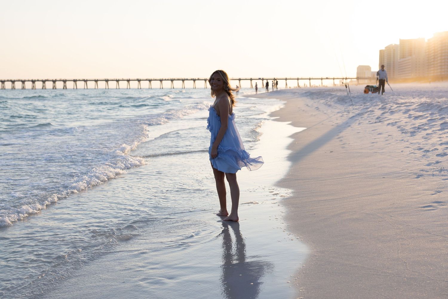 A woman in a blue dress stands barefoot on a sandy beach at sunset with a pier in the background.