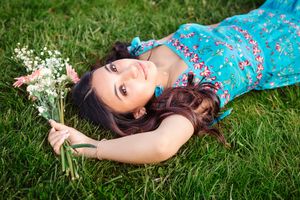 Young woman in blue floral dress lying on green grass holding a bouquet of pink and white flowers.
