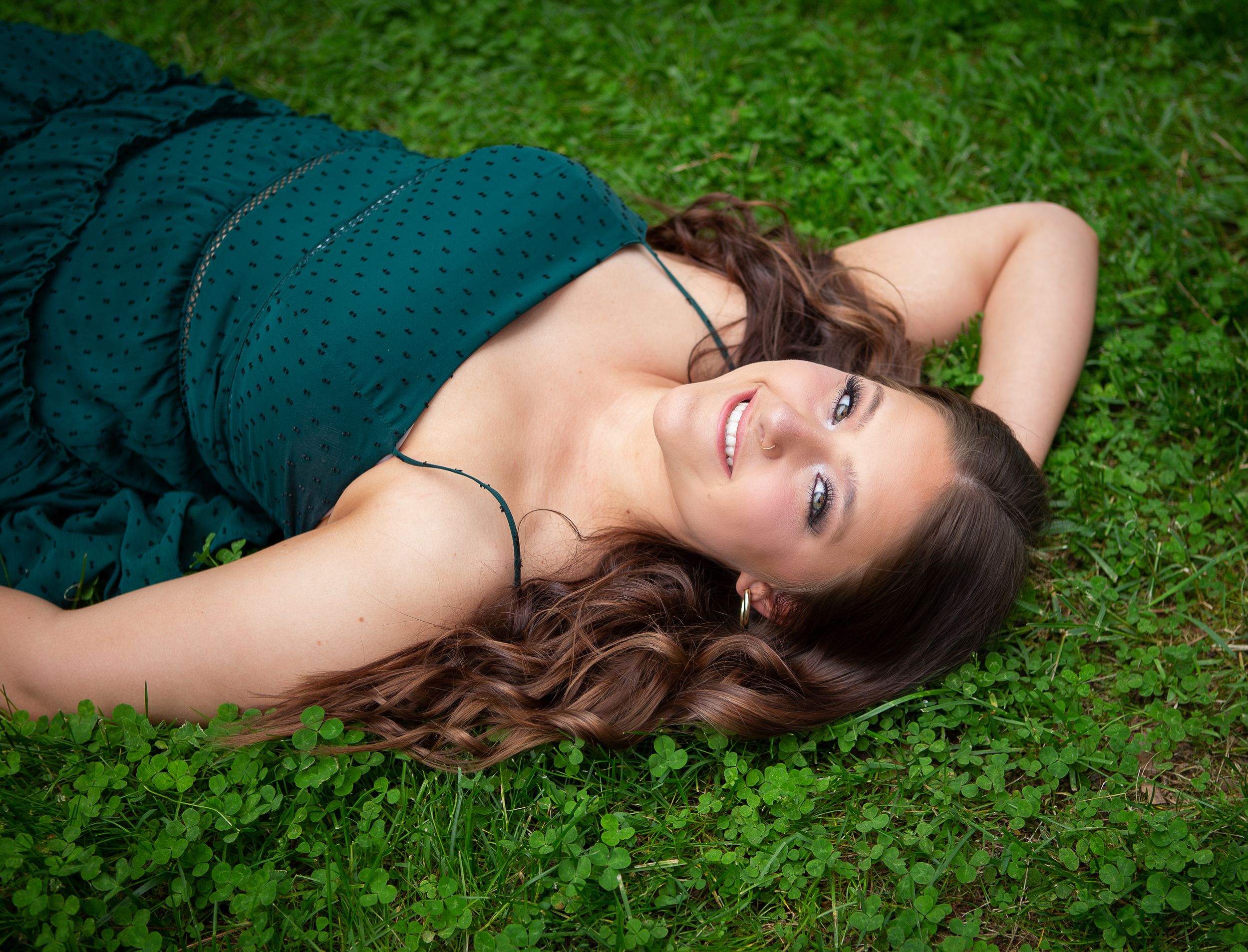 A smiling young woman in a teal dress lying on lush green clover grass with wavy brown hair in Sylva NC.