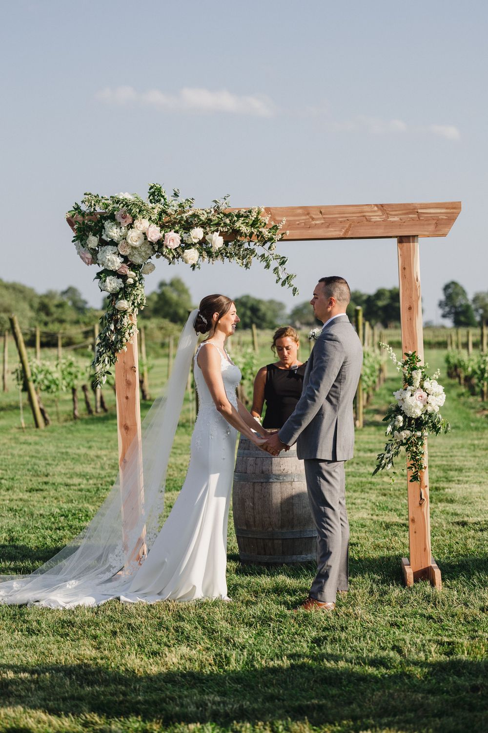 Outdoor wedding ceremony setup with wooden arch and wine barrel floral arrangements in a vineyard.