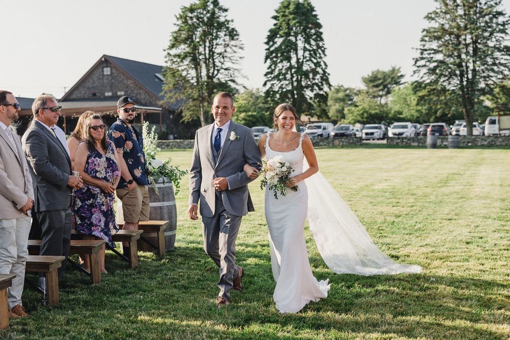 Wedding guests enjoy outdoor reception activities on a sunny day in a grassy field with trees in the background.