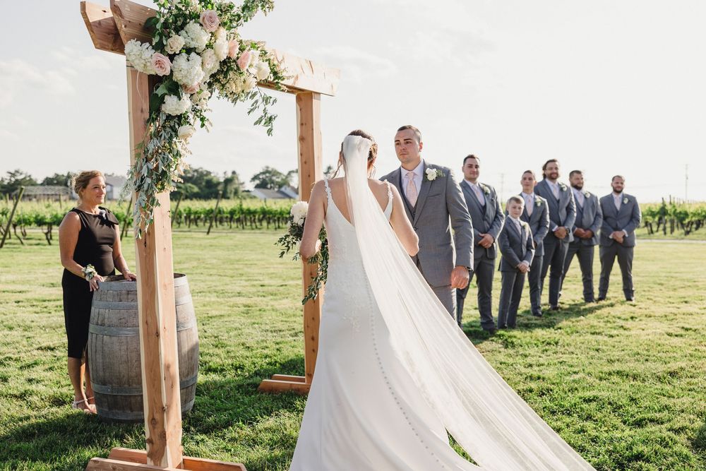 A wedding ceremony at a vineyard with a wooden arch decorated with white flowers and greenery.