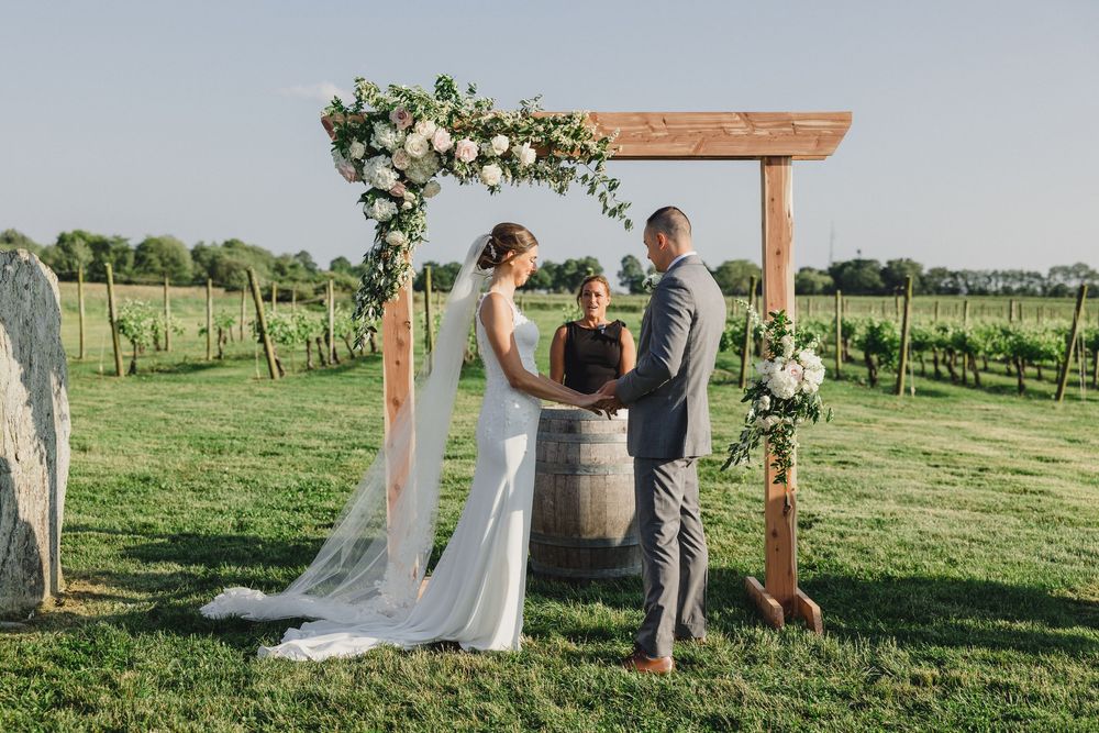 An intimate wedding ceremony takes place beneath a rustic wooden arbor in a scenic vineyard setting.