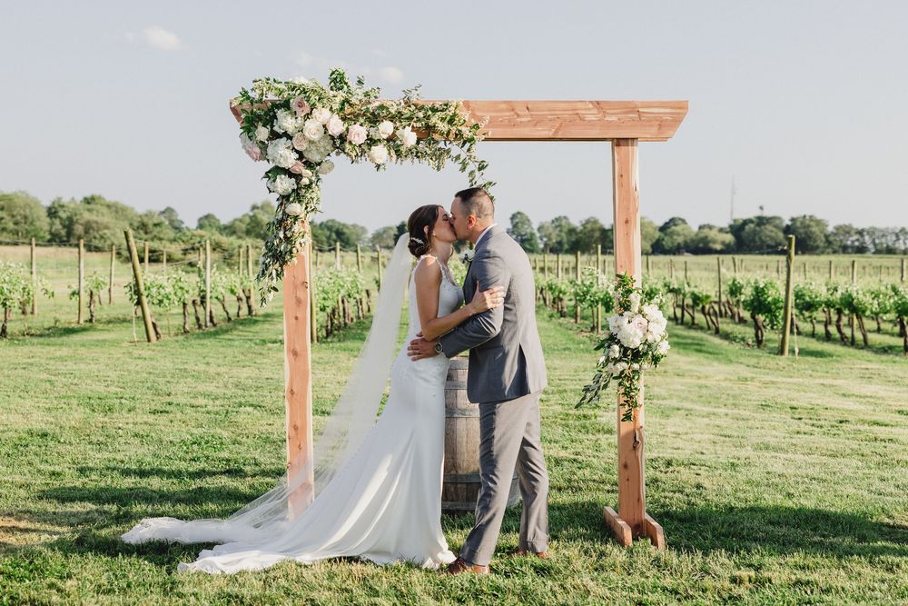 A romantic wedding moment captured under a wooden arch in a sunlit vineyard.