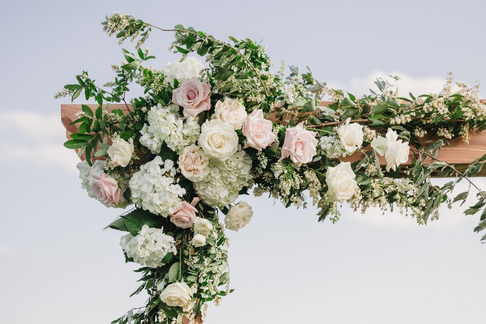 Wedding arch decorated with lush greenery and blush pink and white roses against a cloudy sky backdrop.