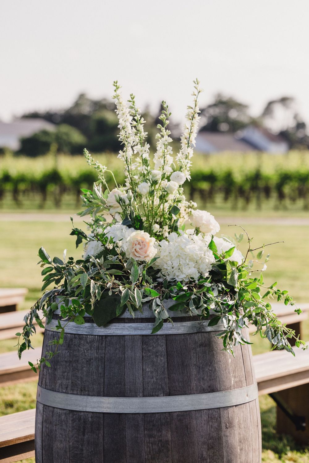 Elegant white floral arrangements with roses and greenery displayed on rustic wine barrels in an outdoor vineyard setting.