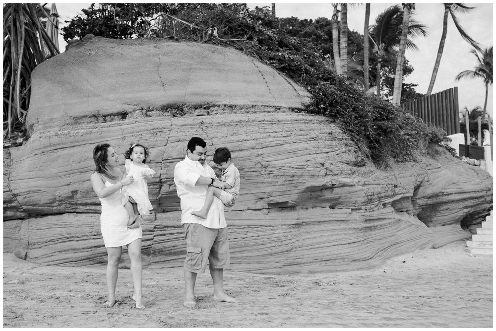 Two couples in white outfits dance together on a beach with rock formations in the background.