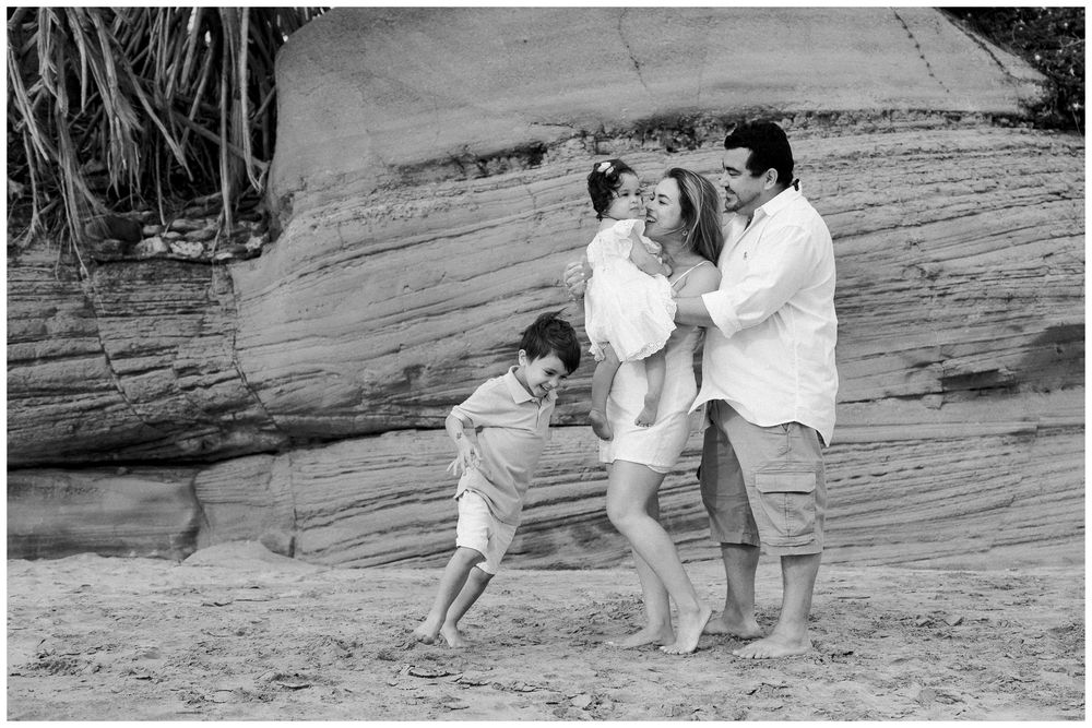 A family enjoys a playful moment together on a rocky beach during a casual outdoor photoshoot.