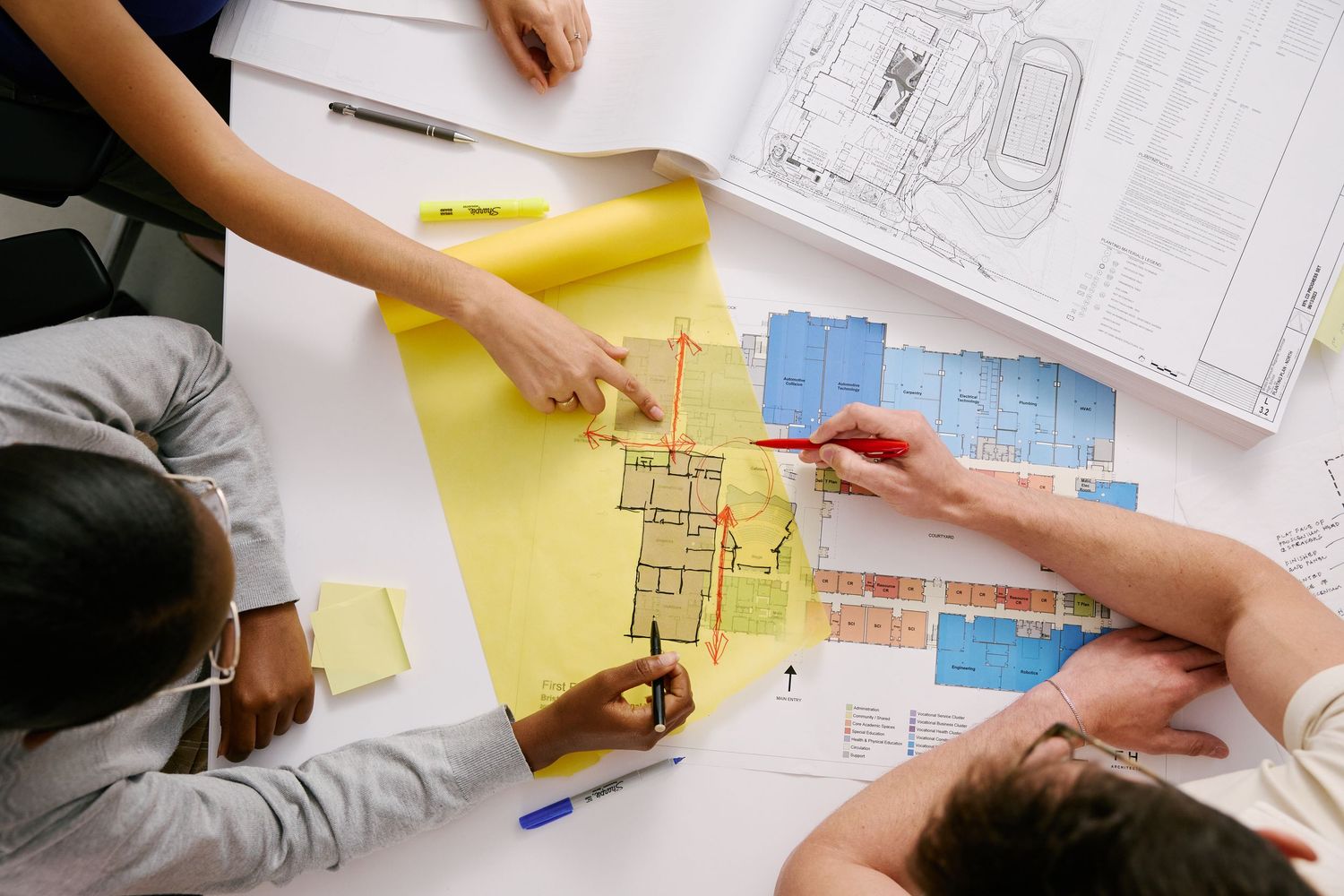 Hands pointing at architectural blueprints and yellow papers spread across a white table during a planning meeting.