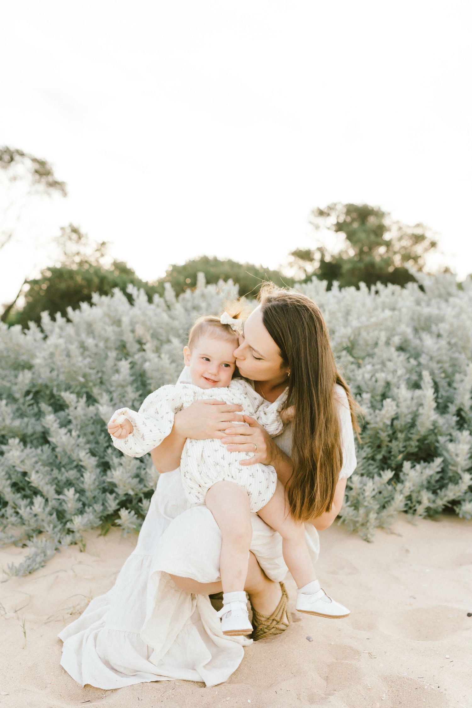 A parent and child in white dresses share tender moments on a sandy path surrounded by silvery desert brush.