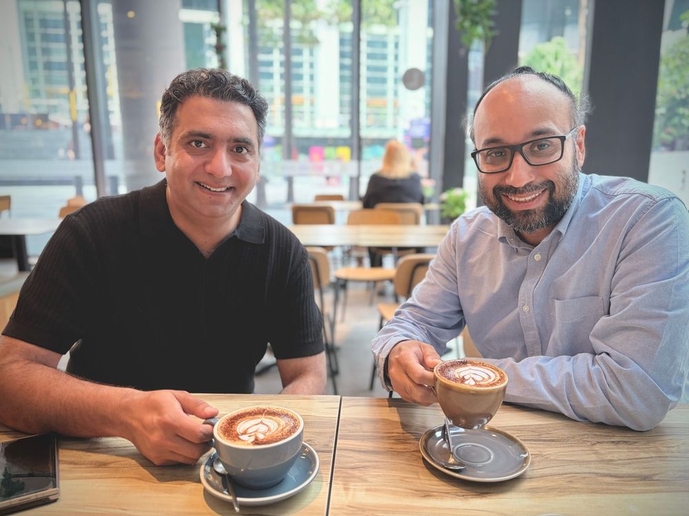 Two men smiling holding latte art coffees at cafe table.