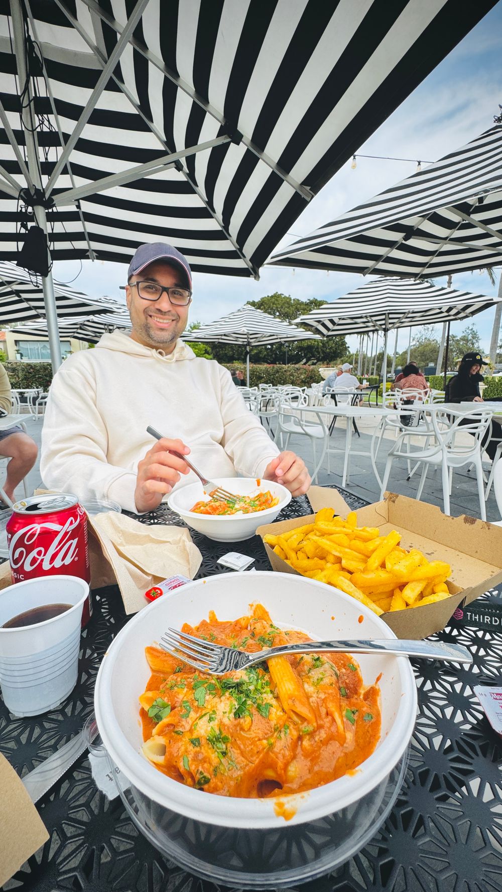 Man smiling while enjoying a meal of pasta and fries at an outdoor cafe with striped umbrellas.