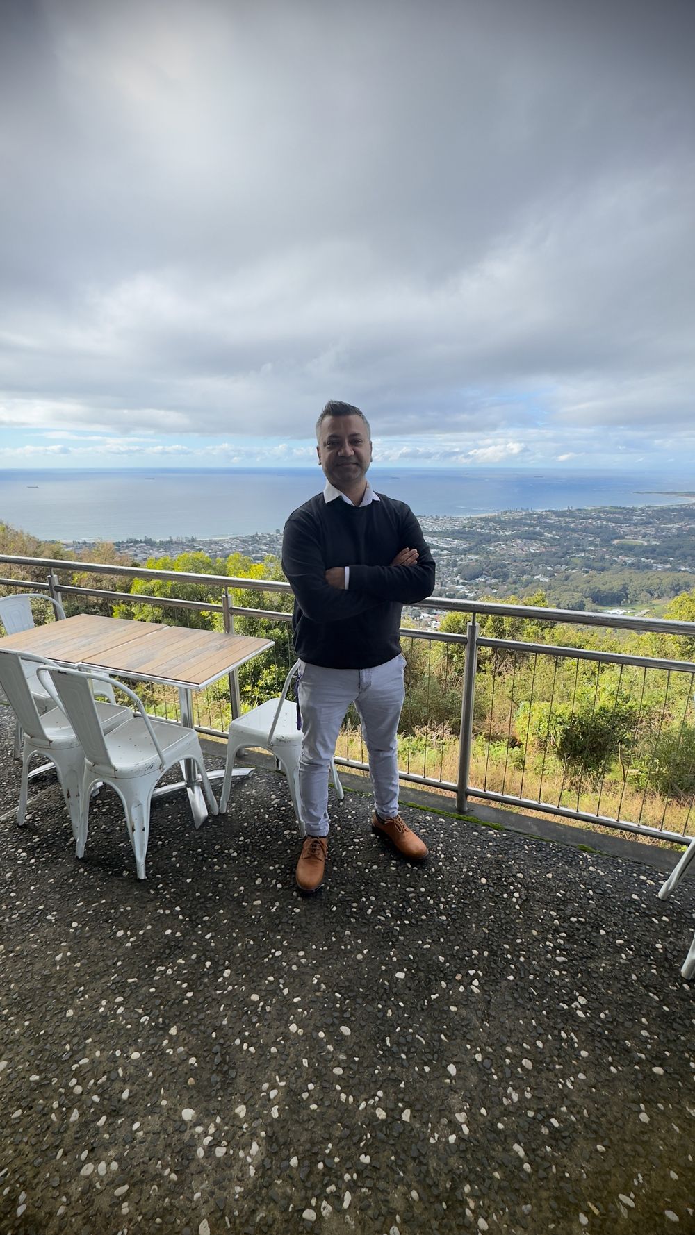 Man in black sweater standing on a scenic hilltop terrace overlooking a coastal town.