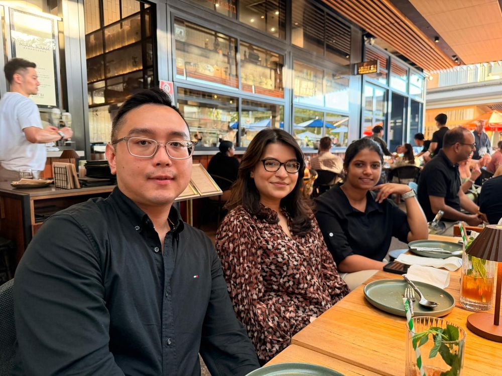 Three colleagues smiling together at a busy restaurant dining table.