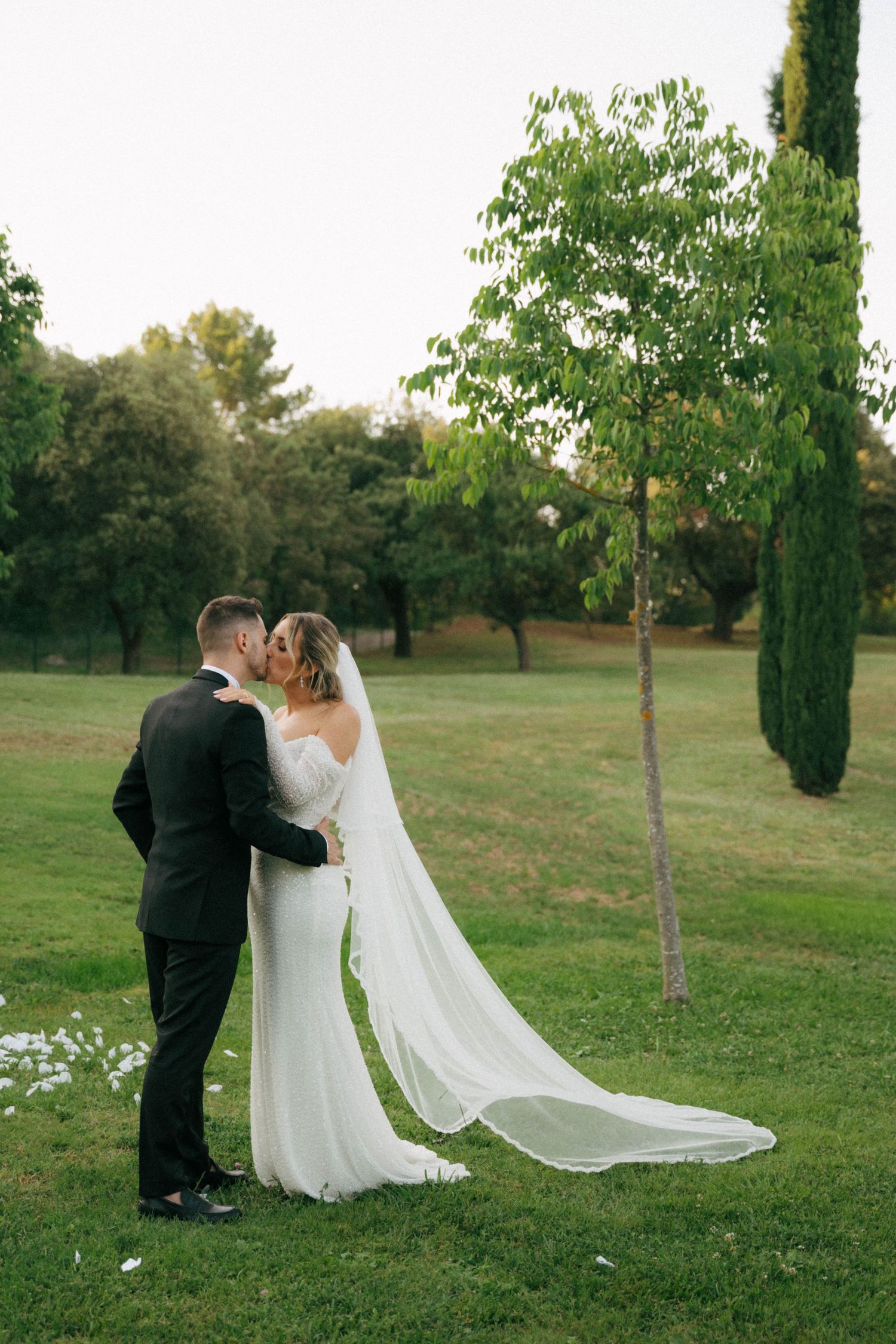 A romantic wedding couple shares an intimate moment in a serene outdoor garden setting with flowing white veil and formal attire.